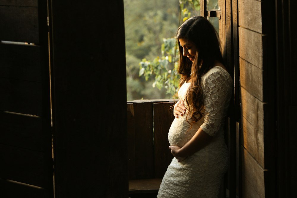 A mother-to-be standing by a sunlit window with her hands on her belly